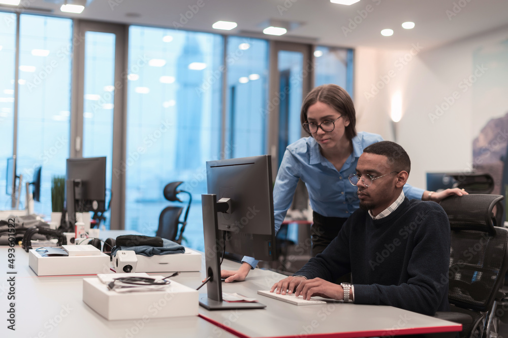 Young smiling woman explaining to serious African American coworker project strategy. Diverse startup coworkers students woman and man talking discussing working in modern office using computer. Young smiling woman explaining to serious African American coworker project strategy. Diverse startup coworkers students woman and man talking discussing working in modern office using computer.