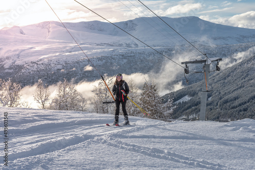 Woman with brown hair smiling, wearing black ski wear, on ski lift moving up, beautiful mountaing range and valley in background with sunlight, horizontal photo