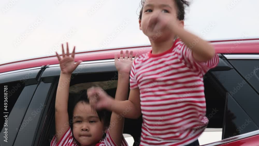 Cute Asian siblings girls smiling and having fun traveling by car and looking out of the car window. Happy family enjoying road trip on summer vacation.