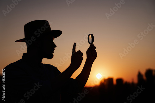 Silhouette of detective male in hat looking with magnifying glass raising index finger gesture. Man searching sales or discounts at sunset using loupe with sun on background and urban city view