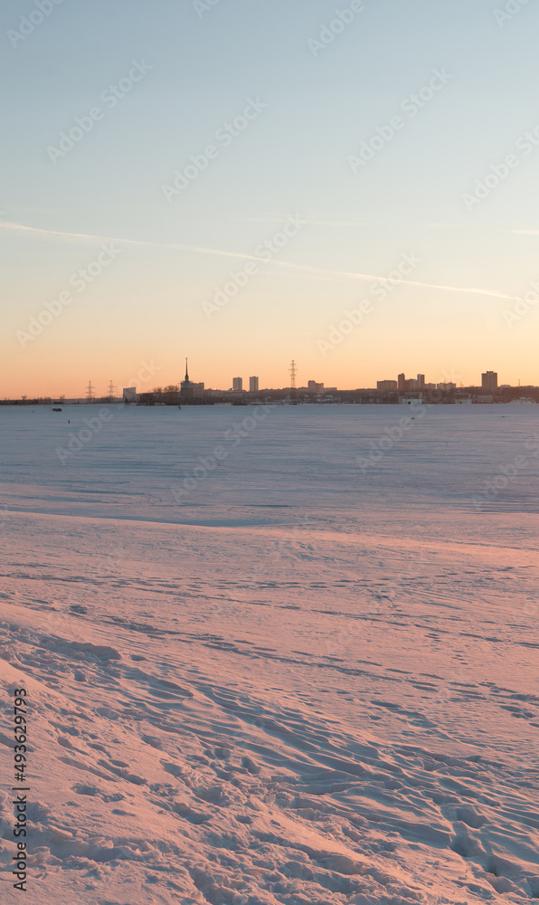 View of the snow-covered river and the city on the far bank at sunset.