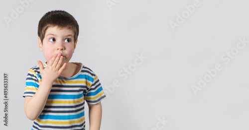A handsome boy of 4 years old in a striped T-shirt covers his mouth with his hand and looks to the side, a banner with a place for text