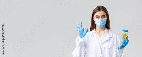 Covid-19, medical research, healthcare workers and quarantine concept. Professional doctor in scrubs, medical mask and gloves, holding patient urine sample and show okay sign, approve, making tests
