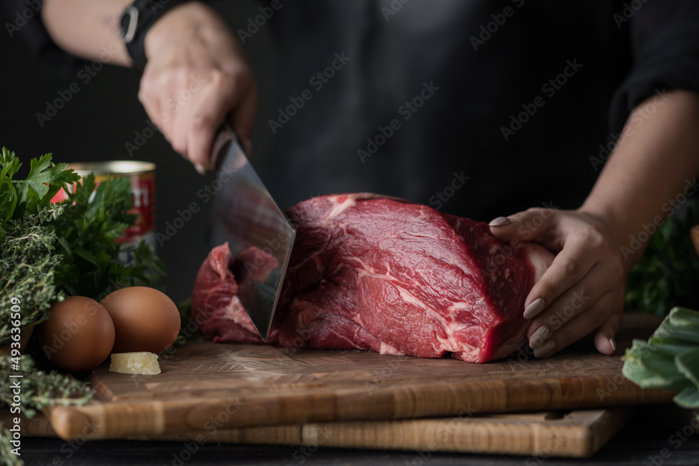 Dark and Moody. Chef in  holds a big knife and cuts into pieces raw meat on a brown wooden cutting board