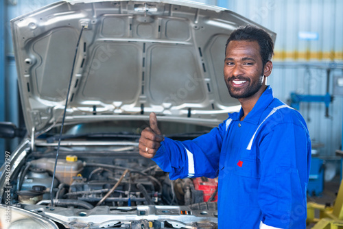 young car mechanic working by showing thumb up by looking at camera at garage - concept of approval, car repair service and professional worker.