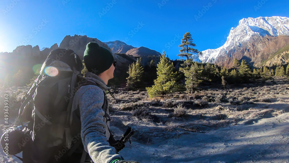 A man taking a selfie while trekking along Annapurna Circuit in Nepal. He is enjoying the view and trek. There is a lush green Himalayan valley around him. Snow caped mountains in the back