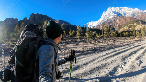 A man taking a selfie while trekking along Annapurna Circuit in Nepal. He is enjoying the view and trek. There is a lush green Himalayan valley around him. Snow caped mountains in the back photo