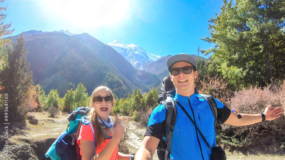 A couple taking a selfie while trekking along Annapurna Circuit in Nepal. They are having fun and making silly faces. There is a lush green Himalayan valley around. Snow caped mountains in the back