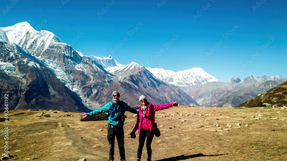 A couple posing with snow caped Annapurna chain behind them, along Annapurna Circuit Trek, Nepal. Clear weather, dry grass, snowy peaks. High altitude, massive mountains. Happiness and fun
