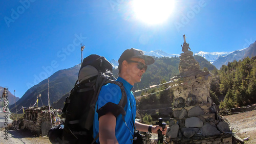 A man taking a selfie while trekking along a small white pagoda, Annapurna Circuit Trek in Nepal. He is enjoying the view and trek. Snow caped mountains in the back. Bright and sunny day.