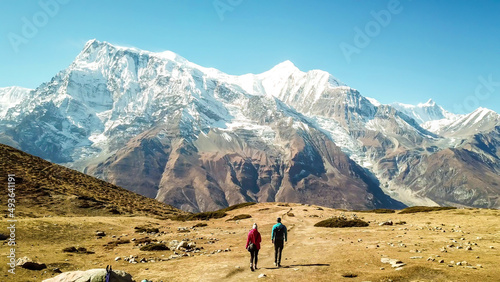 A couple walking on the Annapurna Circuit Trek, Himalayas, Nepal. Snow caped Annapurna chain in the back. Clear weather, dry grass, snowy peaks. High altitude, massive mountains. Freedom and adventure photo
