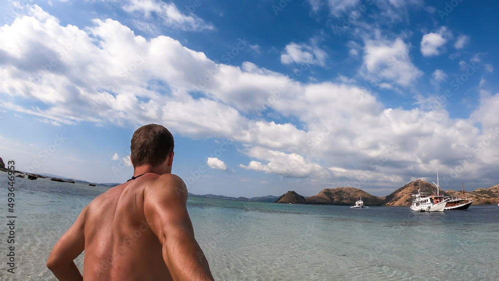 A man walking on a shallow sea water while taking selfies on Kelor Island in Komodo National Park, Indonesia. He is enjoying the moment, having fun while island hopping. There is a boat in the back