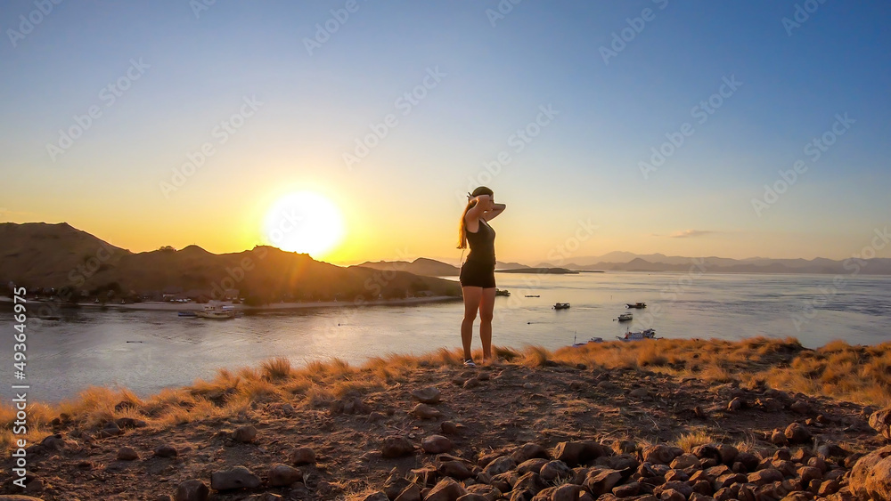 A woman standing on top of a small island, enjoying the morning sun over Komodo National Park, Flores, Indonesia. Golden hour over the islands and sea. Some boats anchored to the bay.