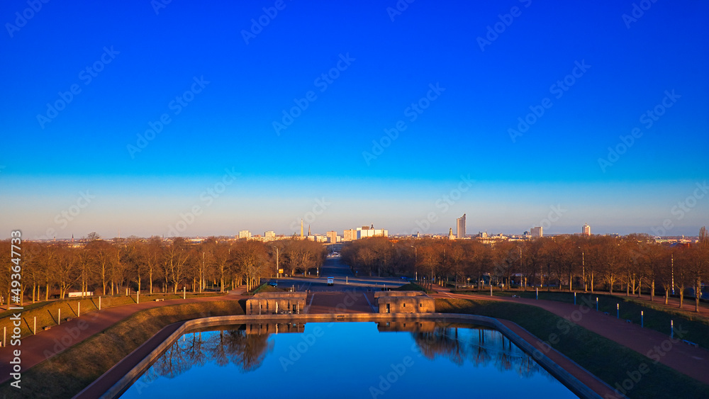 Fototapeta premium Panorama-Blick vom Völkerschlachtdenkmal über den See der Tränen auf die Skyline von Leipzig bei Sonnenaufgang, weite Sichtachse und monumentale Parkanlage, Leipzig, Sachsen, Deutschland