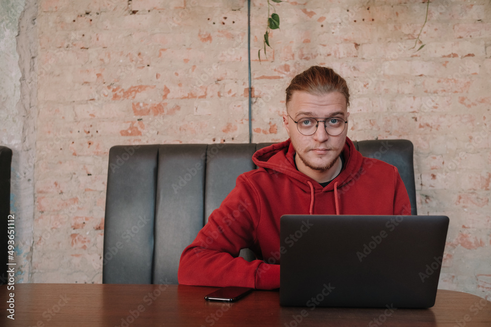 Portrait of happy male freelancer in optical eyewear for vision correction smiling at camera during break from web working online, cheerful hipster blogger sitting in cafe with mockup laptop