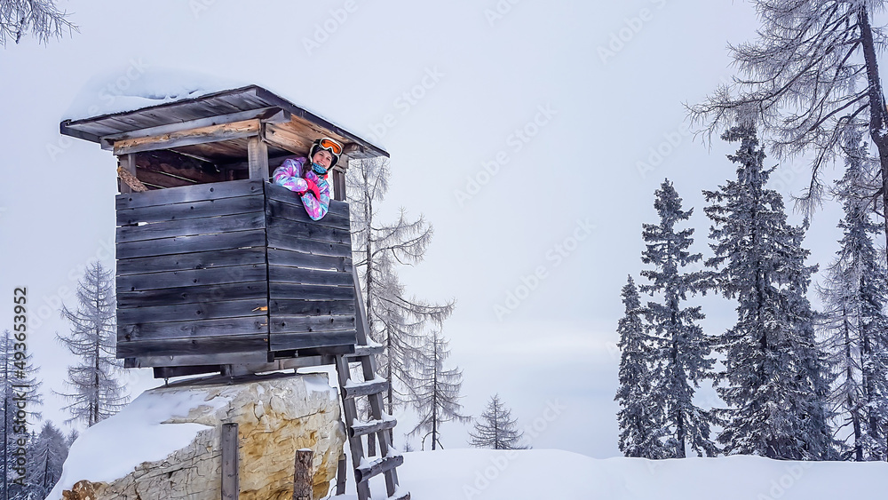Girl in a skiing outfit and helm standing inside of a hunting pulpit, and leaning through the window. The ski slope is wreathed with fog. The visibility is very low. Girl is happy and enjoying herself Girl in a skiing outfit and helm standing inside of a hunting pulpit, and leaning through the window. The ski slope is wreathed with fog. The visibility is very low. Girl is happy and enjoying herself