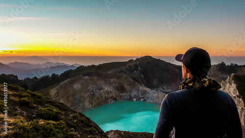 A man watching the sunrise over the Kelimutu volcanic crater lakes in Moni, Flores, Indonesia. Skyline is orange, sun slowly rising. Man is relaxed and calm, enjoying the view on turquoise lake photo