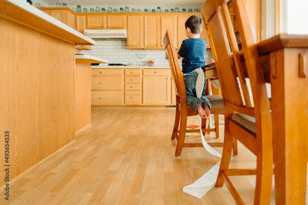 A boy kneels in a dining room chair with a long trail of toilet paper coming out the back of his pants