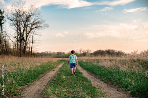 A young boy walks down a two track path between a grass field at sunset
