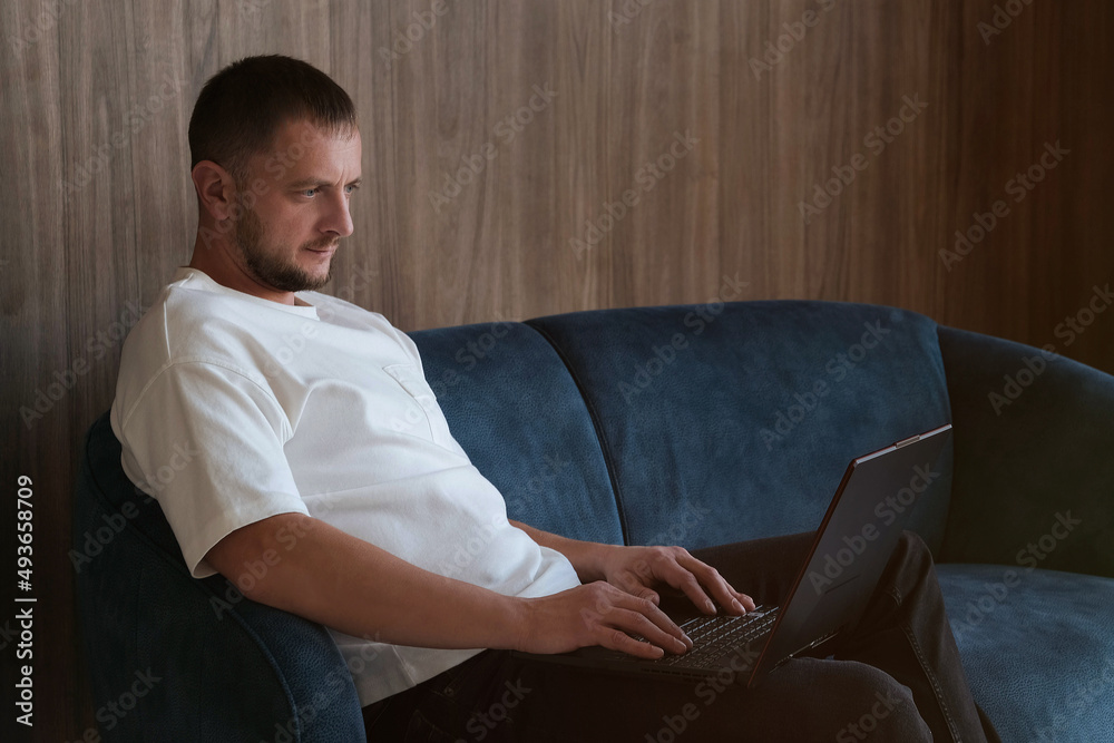 A focused man studying or working on a laptop, sitting on the couch in the living room, a serious freelancer, looking at computer notes, doing research at home. The concept of remote freelance work. A focused man studying or working on a laptop, sitting on the couch in the living room, a serious freelancer, looking at computer notes, doing research at home. The concept of remote freelance work.