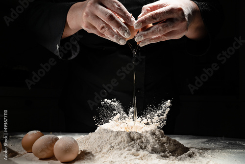 Billede på lærred woman breaks the egg into flour to knead the dough on black background