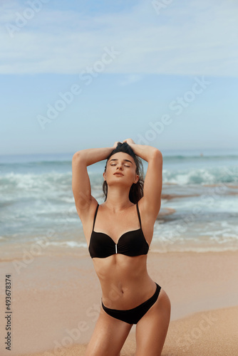 Portrait of pretty beautiful fashion woman in black bikini posing in summer near the sea and blue sky. Young Girl with tanned sexy body in swimwear with copy space. Summer lifestyle. Vacation