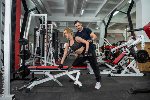 Fit female client working out with personal male instructor with heavy dumbbells at the gym