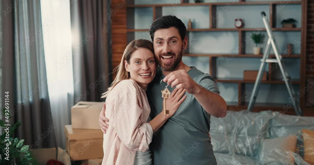 Portrait of loving caucasian couple hugging while holding keys from new home, standing in spacious living room and looking at camera Portrait of loving caucasian couple hugging while holding keys from new home, standing in spacious living room and looking at camera