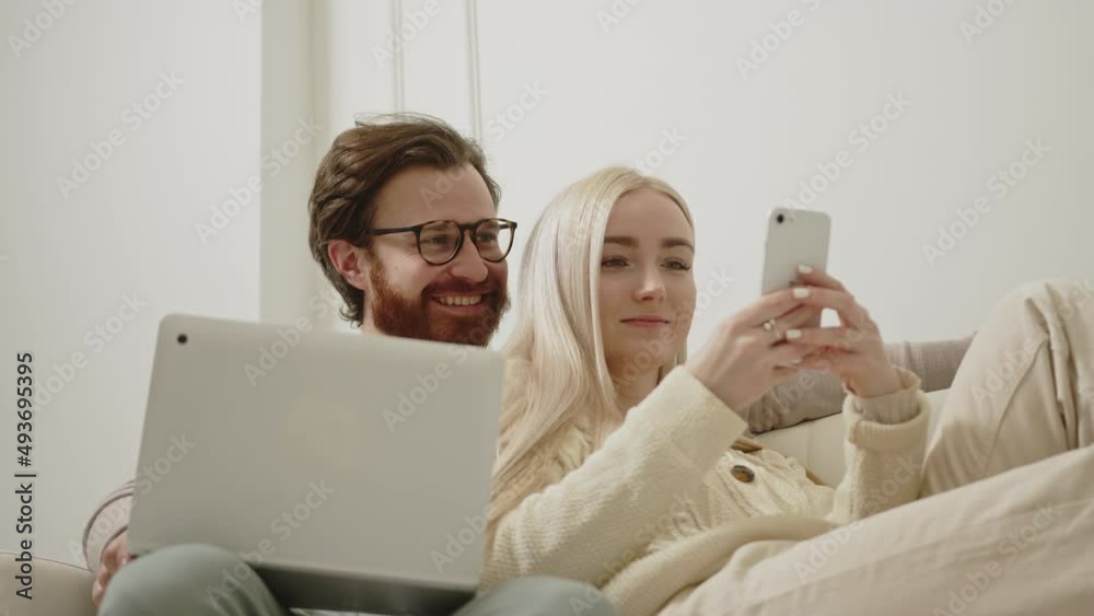portrait of satisfied cheerful millennial couple laughing at a video found in the internet and sitting on white sofa with laptop on knees. High quality 4k footage
