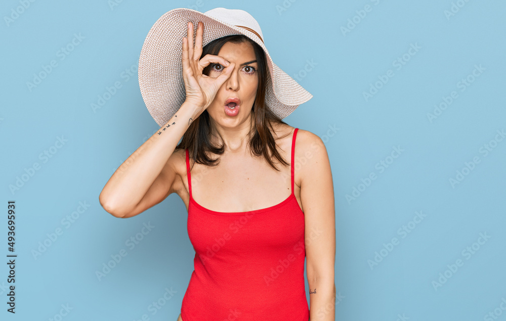 Beautiful brunette woman wearing swimsuit and summer hat doing ok gesture shocked with surprised face, eye looking through fingers. unbelieving expression. Beautiful brunette woman wearing swimsuit and summer hat doing ok gesture shocked with surprised face, eye looking through fingers. unbelieving expression.