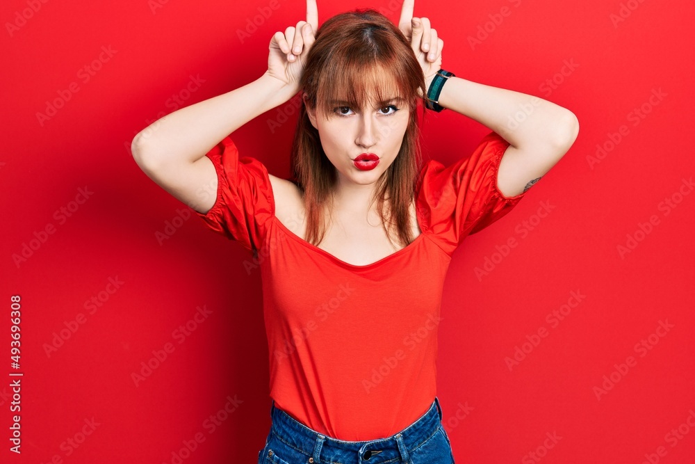 Redhead young woman wearing casual red t shirt doing funny gesture with finger over head as bull horns Redhead young woman wearing casual red t shirt doing funny gesture with finger over head as bull horns