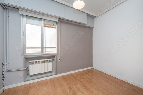 Empty room with oak wooden floor, gray walls and aluminum radiator with a window that communicates with a closed terrace