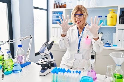 Middle age blonde woman working at scientist laboratory showing and pointing up with fingers number ten while smiling confident and happy.