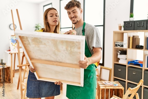 Young hispanic artist couple smiling happy holding canvas at art studio.