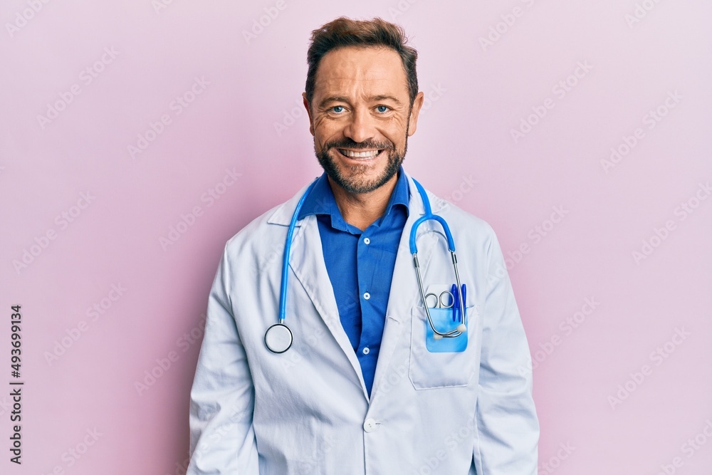 Middle age man wearing doctor uniform and stethoscope looking positive and happy standing and smiling with a confident smile showing teeth Middle age man wearing doctor uniform and stethoscope looking positive and happy standing and smiling with a confident smile showing teeth
