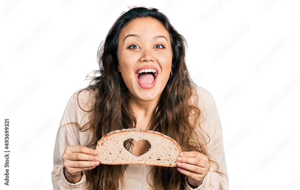 Young hispanic girl holding bread with heart symbol celebrating crazy and amazed for success with open eyes screaming excited. Young hispanic girl holding bread with heart symbol celebrating crazy and amazed for success with open eyes screaming excited.