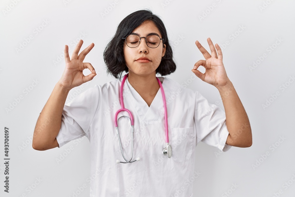 Young hispanic doctor woman wearing stethoscope over isolated background relax and smiling with eyes closed doing meditation gesture with fingers. yoga concept. Young hispanic doctor woman wearing stethoscope over isolated background relax and smiling with eyes closed doing meditation gesture with fingers. yoga concept.