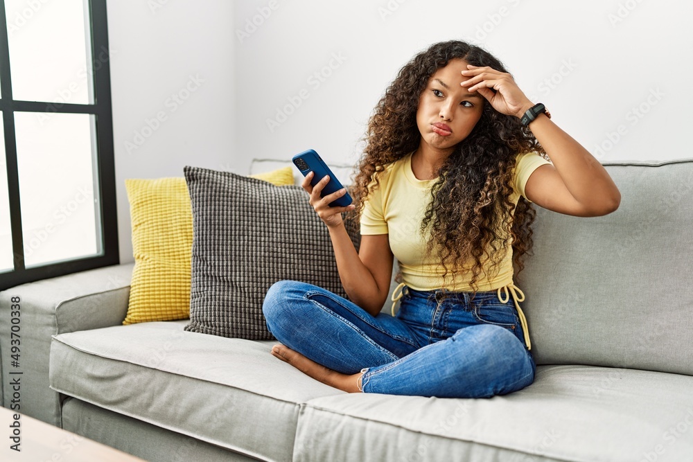 Beautiful hispanic woman sitting on the sofa at home using smartphone worried and stressed about a problem with hand on forehead, nervous and anxious for crisis Beautiful hispanic woman sitting on the sofa at home using smartphone worried and stressed about a problem with hand on forehead, nervous and anxious for crisis
