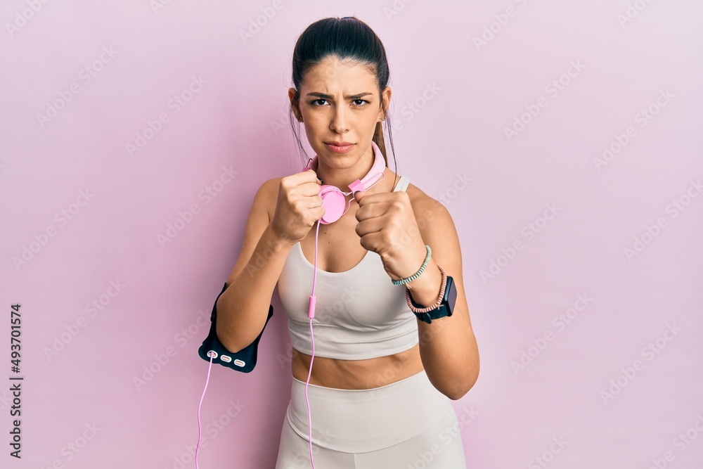Young hispanic woman wearing gym clothes and using headphones ready to fight with fist defense gesture, angry and upset face, afraid of problem