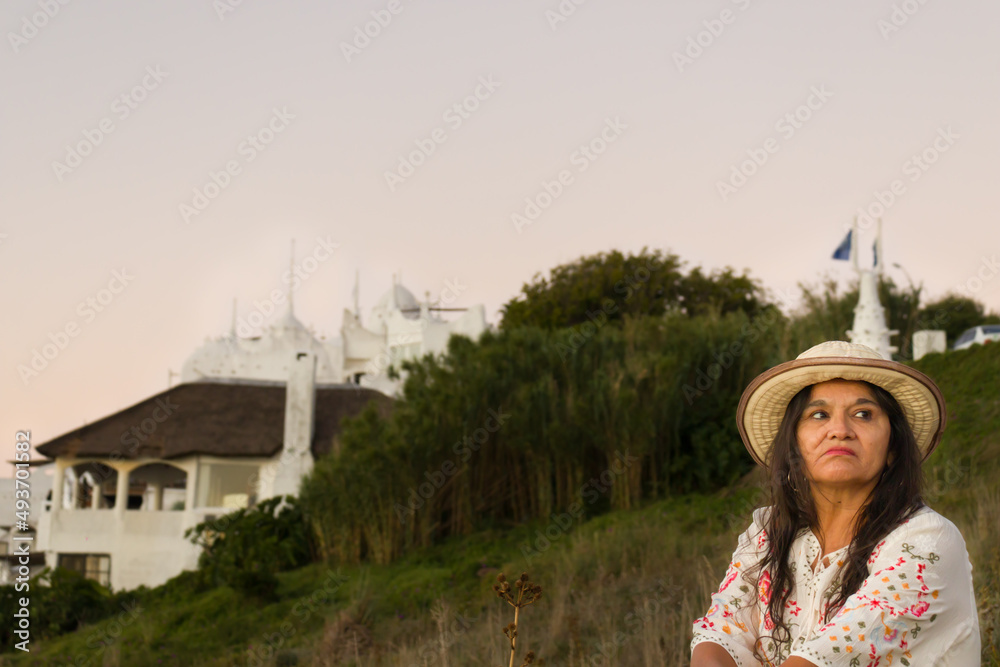 mujer latina. Señora, adulta con gafas de sol, sombrero, vestida de blanco mirando la puesta de sol en una colina con un fondo blanco. Afuera, al aire libre.
