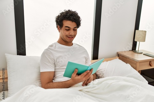 Young hispanic man reading book sitting on the bed at bedroom.