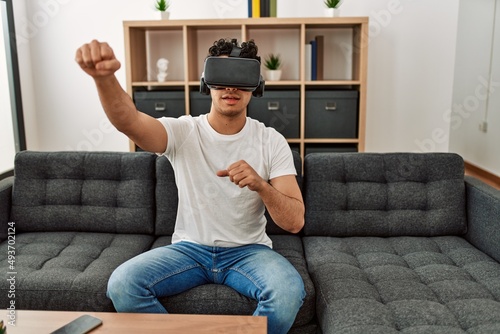 Young hispanic man playing with virtual reality glasses sitting on the sofa at home. photo