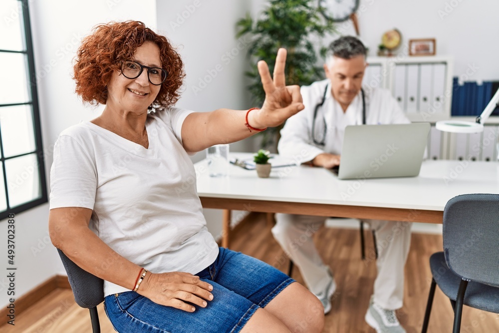 Senior woman sitting at doctor appointment smiling looking to the camera showing fingers doing victory sign. number two. Senior woman sitting at doctor appointment smiling looking to the camera showing fingers doing victory sign. number two.