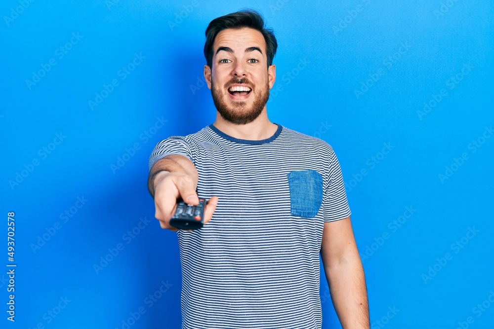 Handsome caucasian man with beard holding television remote control celebrating crazy and amazed for success with open eyes screaming excited. Handsome caucasian man with beard holding television remote control celebrating crazy and amazed for success with open eyes screaming excited.