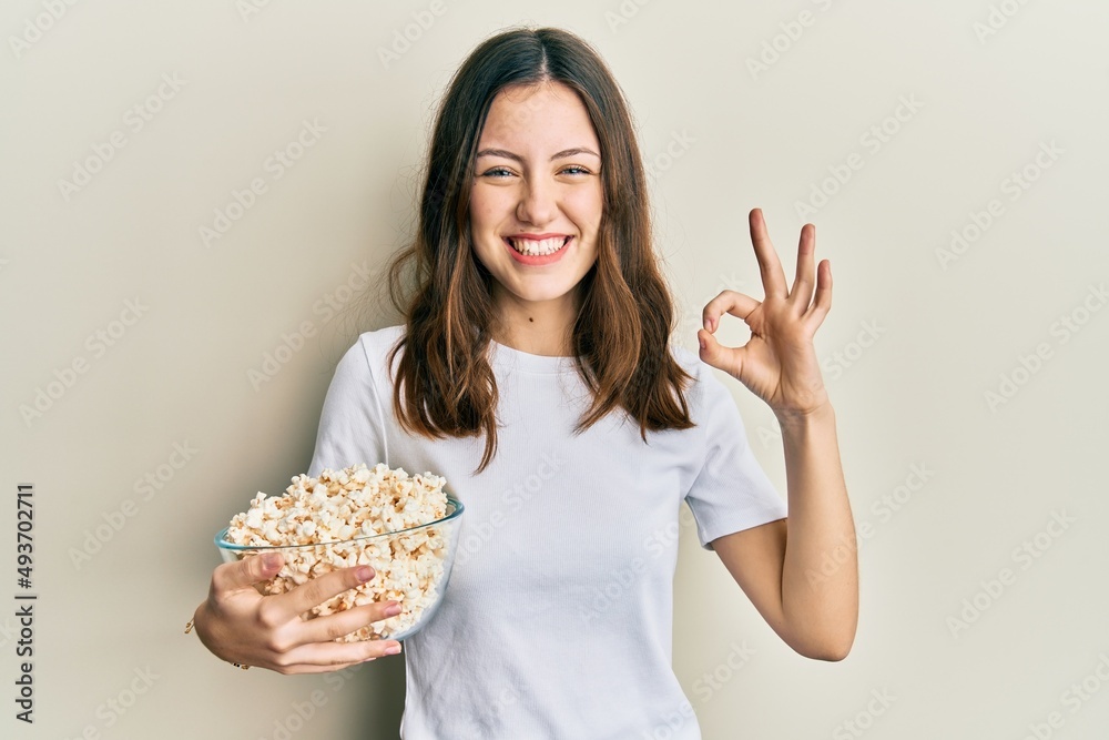 Young brunette woman eating popcorn doing ok sign with fingers, smiling friendly gesturing excellent symbol Young brunette woman eating popcorn doing ok sign with fingers, smiling friendly gesturing excellent symbol