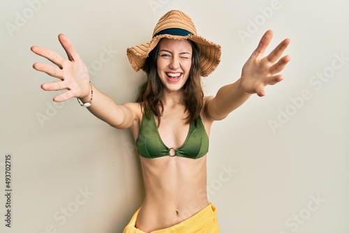 Young brunette woman wearing bikini and hat with open arms hugging winking looking at the camera with sexy expression, cheerful and happy face.