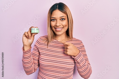 Beautiful hispanic woman holding cosmetic moisturizer cream smiling happy pointing with hand and finger