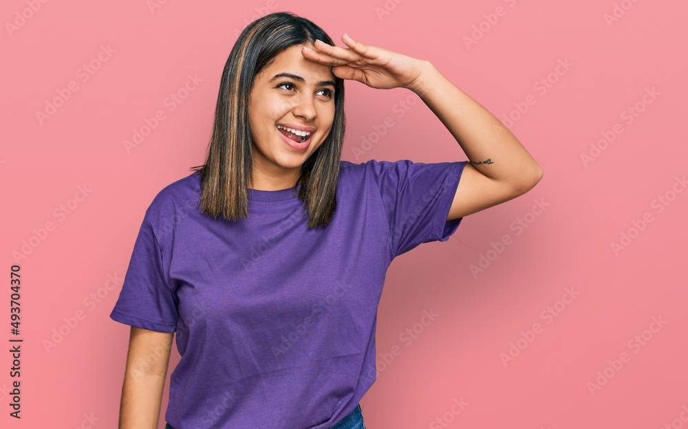 Young hispanic girl wearing casual purple t shirt very happy and smiling looking far away with hand over head. searching concept. Young hispanic girl wearing casual purple t shirt very happy and smiling looking far away with hand over head. searching concept.