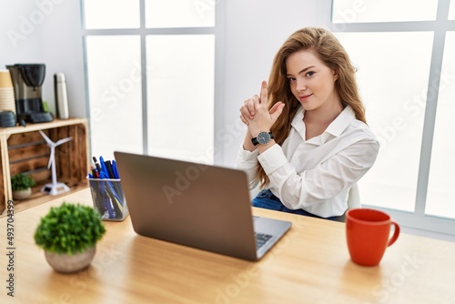 Young caucasian woman working at the office using computer laptop holding symbolic gun with hand gesture, playing killing shooting weapons, angry face