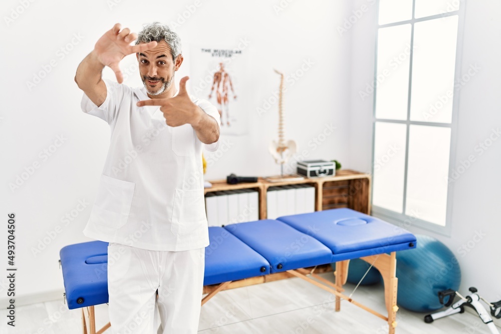 Middle age hispanic therapist man working at pain recovery clinic smiling making frame with hands and fingers with happy face. creativity and photography concept.